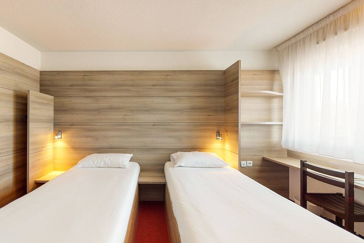 Habitación de hotel con dos camas individuales, cabecero de madera, lámparas de noche, un escritorio y una silla.
