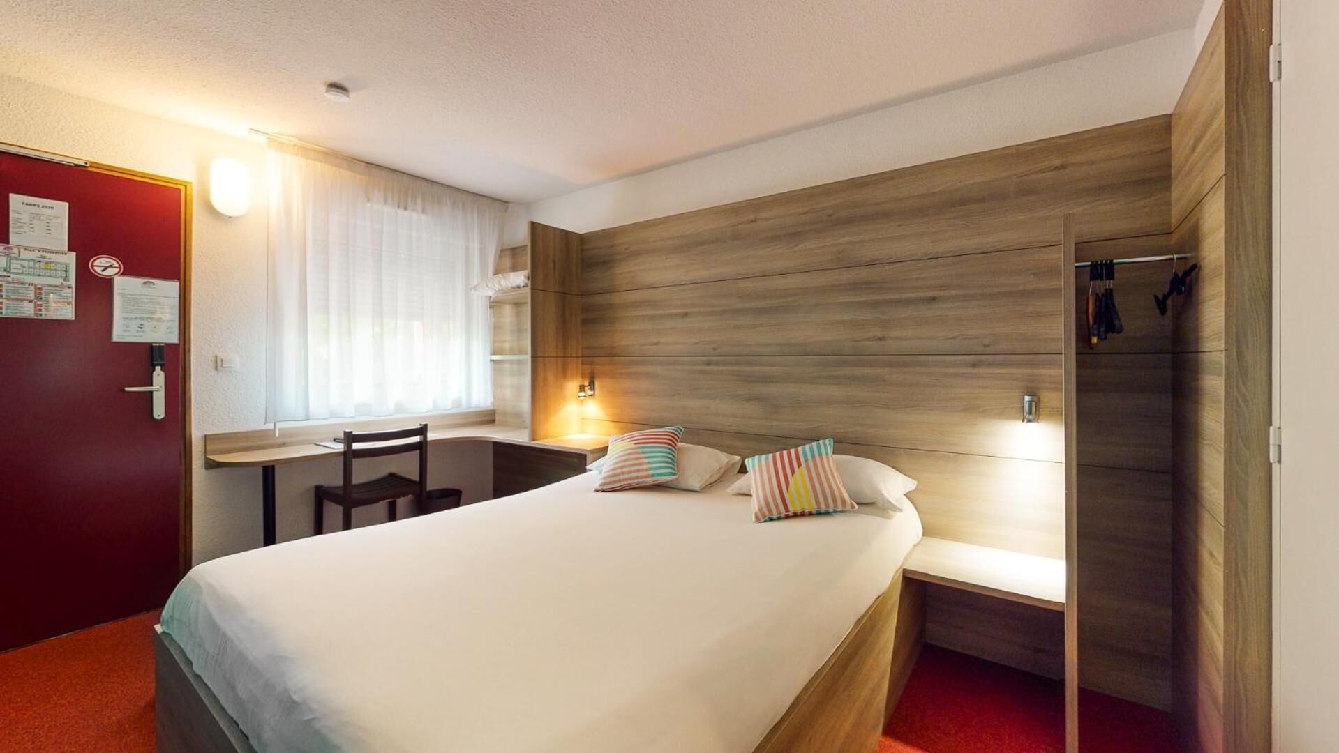 A hotel room with a bed, a desk, and a chair. There is a window with curtains, a wooden headboard, and a red door.