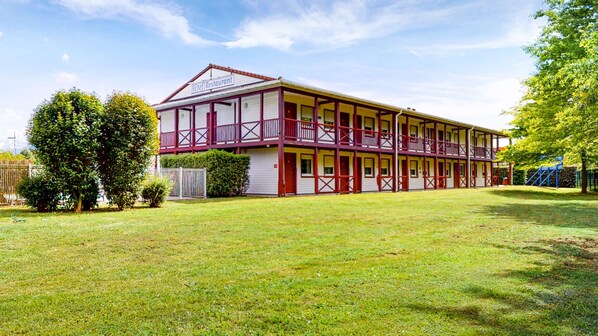 A two-story hotel with a red and white facade, multiple balconies, and a grassy courtyard.
