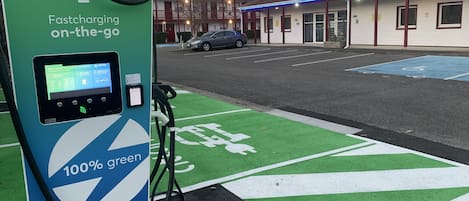 Una estación de carga rápida con una paleta de colores verde y azul, identificada como 100% ecológica, y un edificio con techo rojo al fondo.