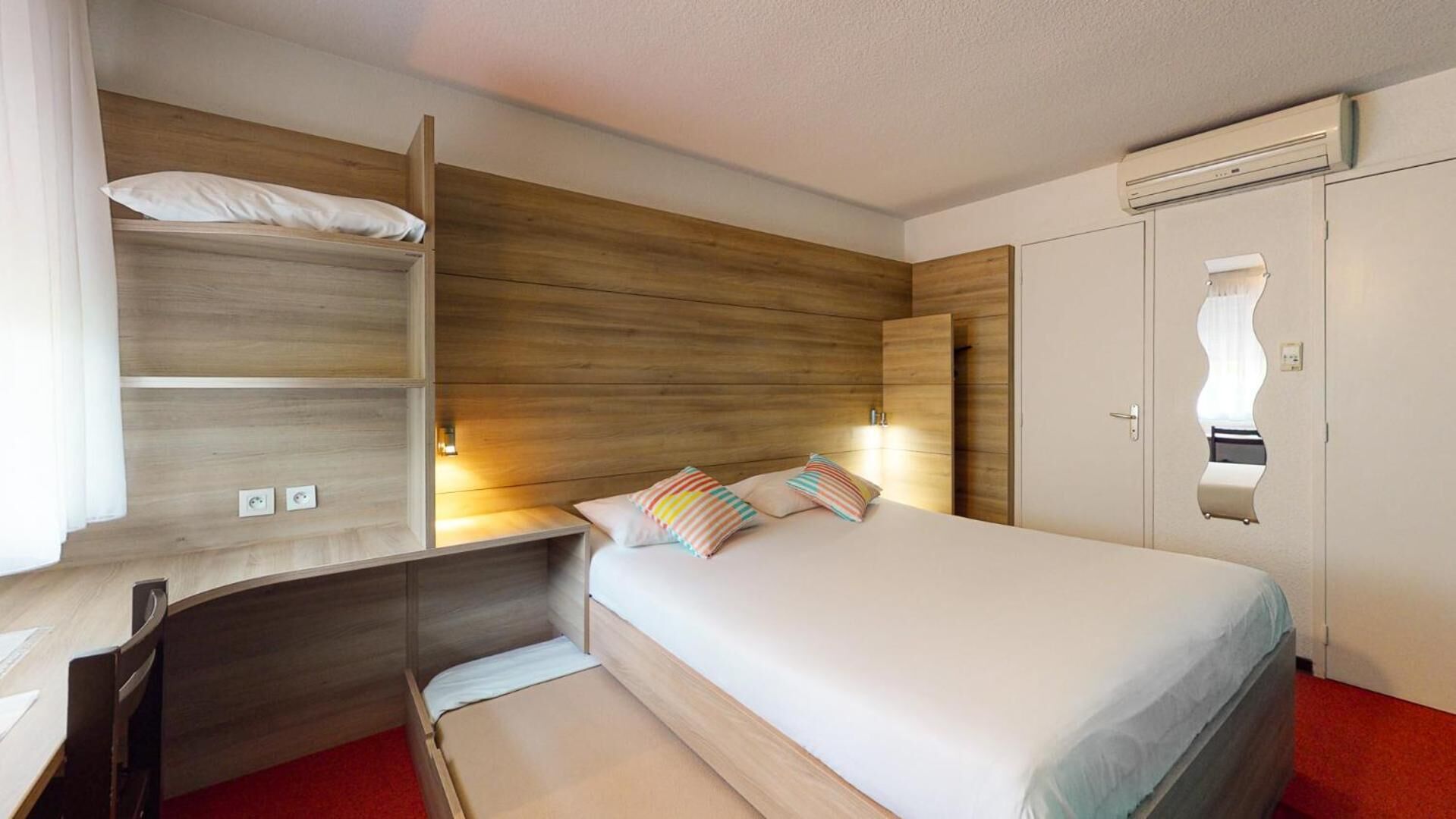 A hotel room with a bed, a wooden headboard, a wardrobe, and a mirror with a wavy design.