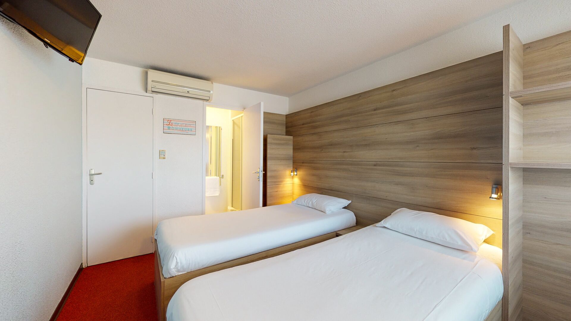 Een hotelkamer met twee enkele bedden, een rood tapijt, een houten hoofdbord en een airconditioning.