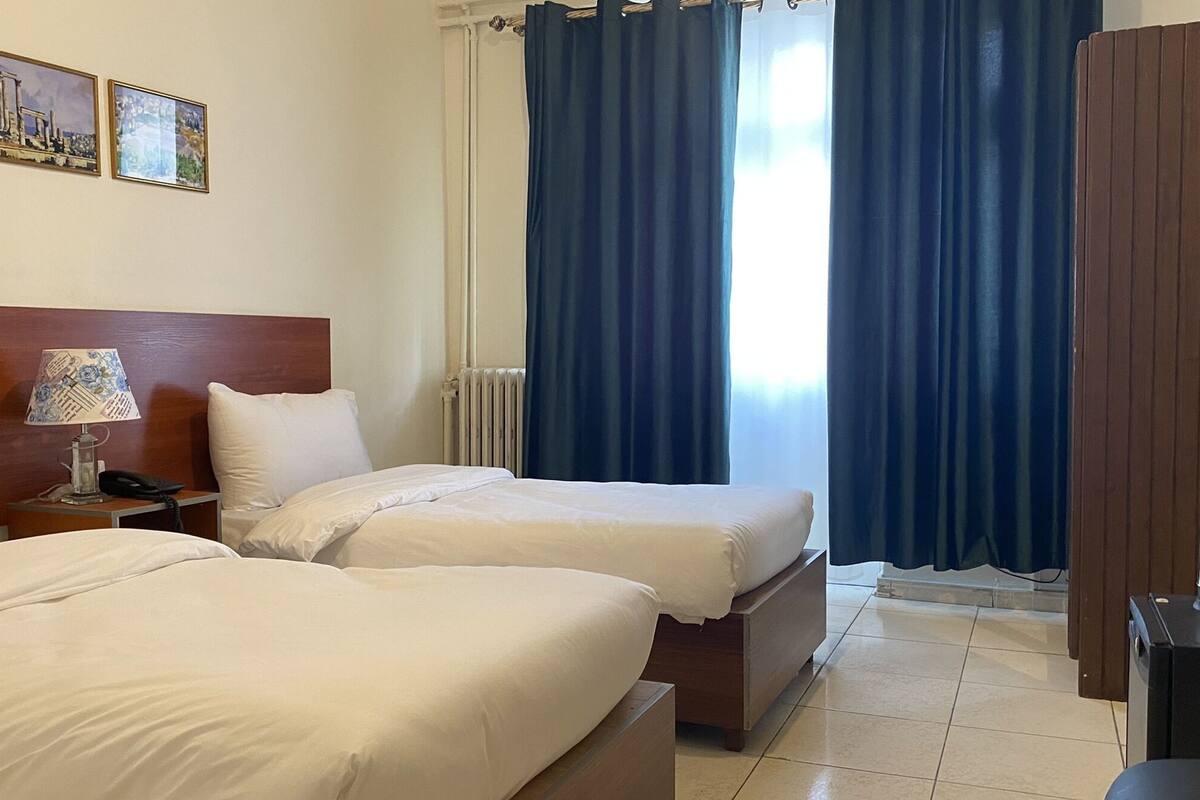 A hotel room with two beds, a wooden headboard, a nightstand with a lamp, a radiator, and a window with blue curtains.