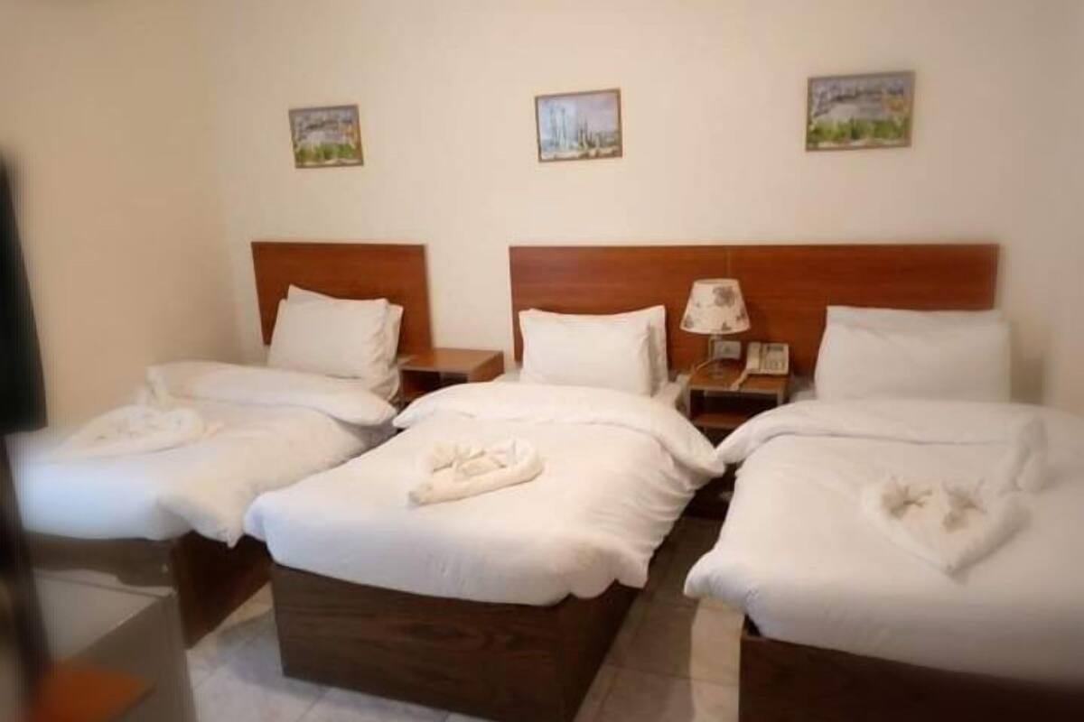 A hotel room with three single beds, wooden headboards, white linens, and a nightstand with a lamp.