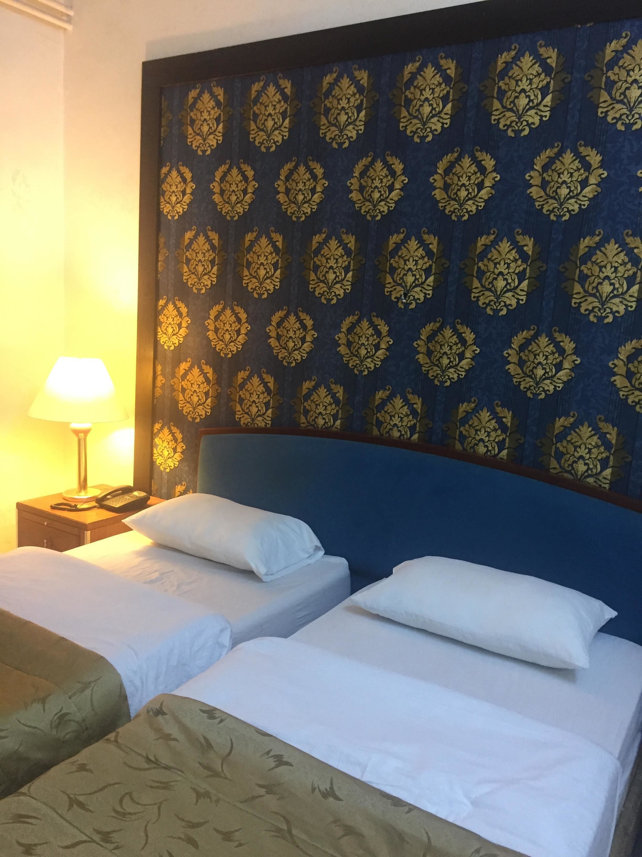 Une chambre d’hôtel avec deux lits, une tête de lit bleue ornée d’un motif doré, du linge de lit blanc et une lampe de chevet.