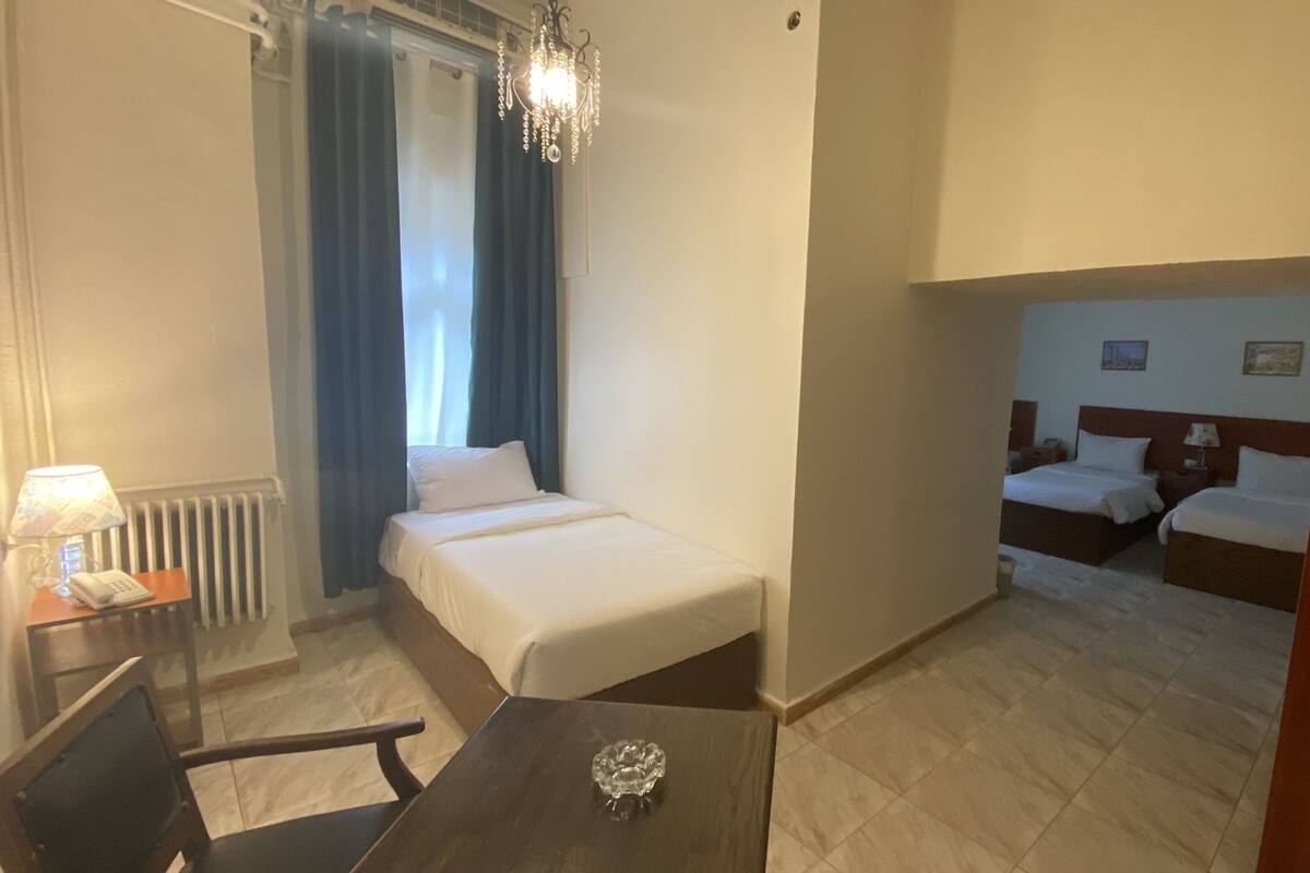 A hotel room with a single bed, a small table with a lamp, a chair, a radiator, and a chandelier.
