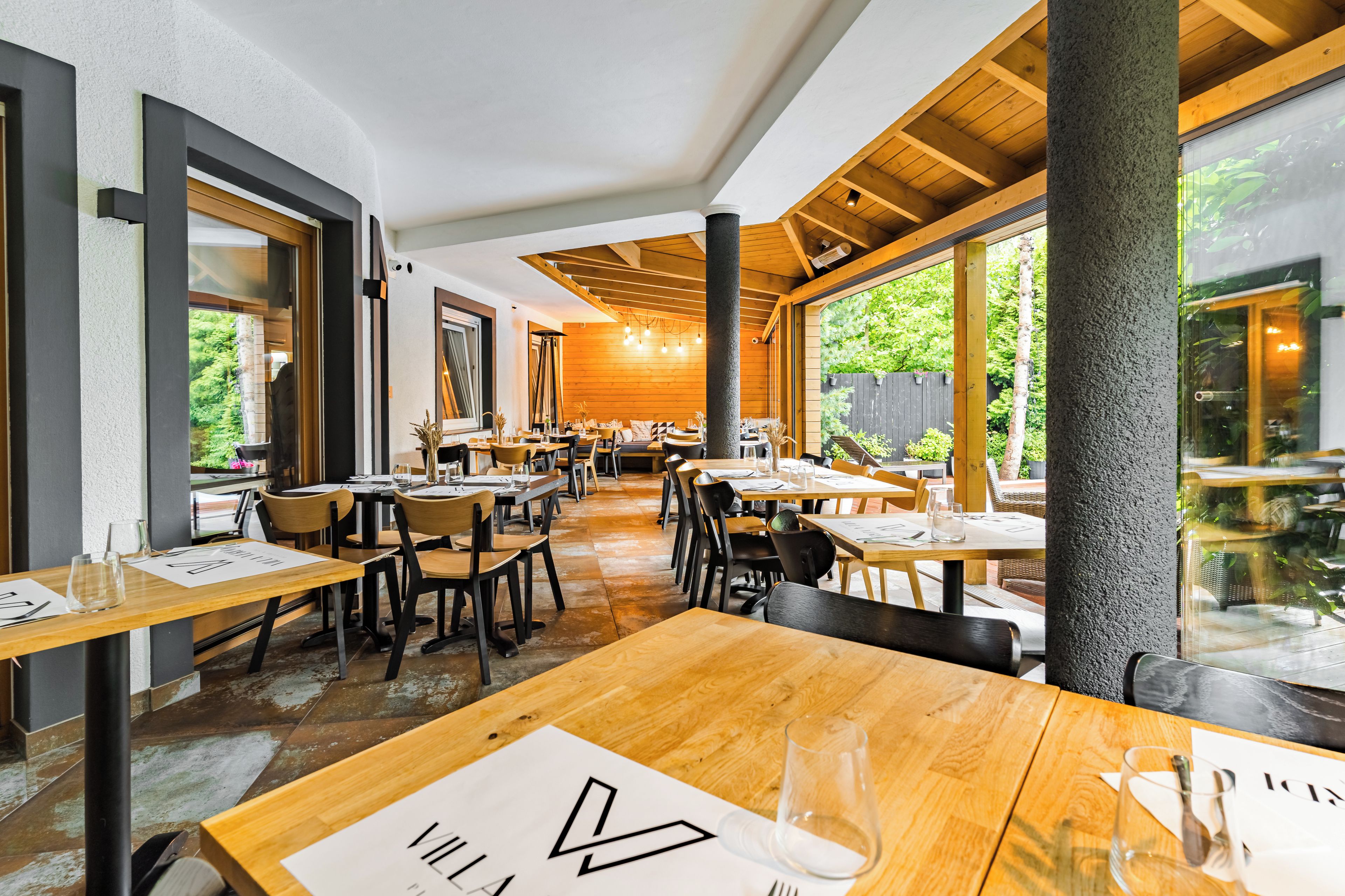 A restaurant interior with wooden tables and chairs, large windows, and a view of greenery outside.