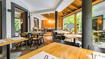 A restaurant interior with wooden tables and chairs, large windows, and a view of greenery outside.