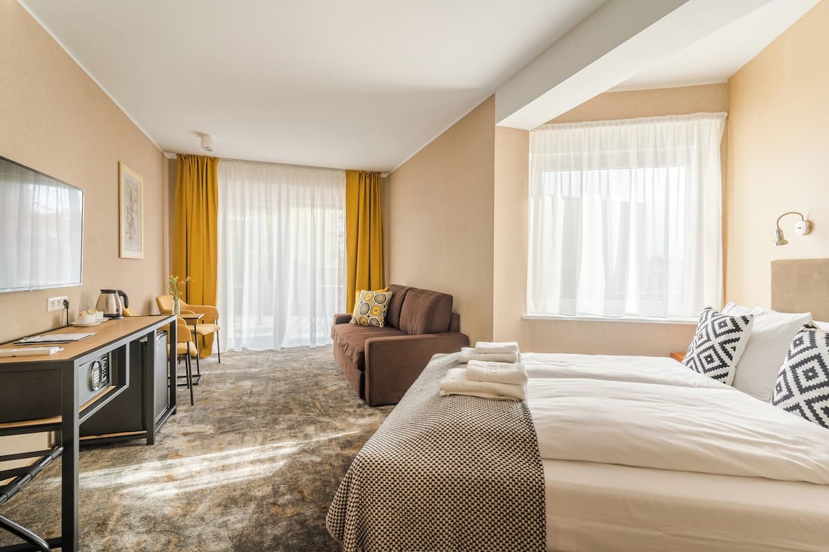 Une chambre d’hôtel avec un lit, un bureau, une chaise et un canapé. Il y a deux fenêtres avec des rideaux.