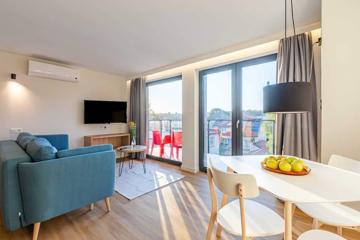 Un salon moderne avec un canapé, un téléviseur, une table à manger avec des chaises et un balcon agrémenté de chaises rouges.
