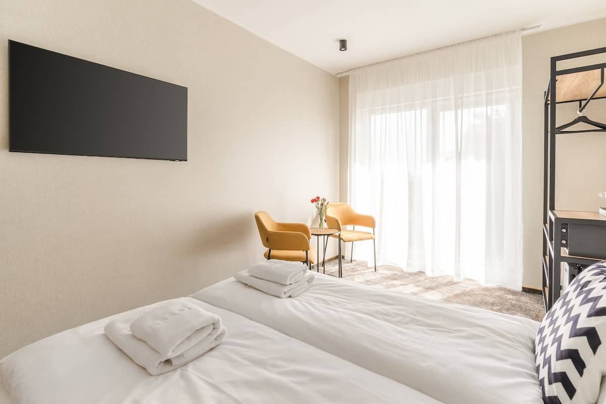 Une chambre d’hôtel avec deux lits, un bureau, une chaise et une grande fenêtre avec des rideaux légers.