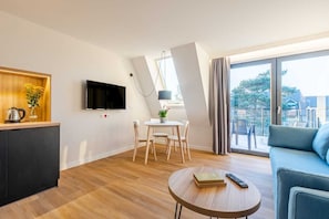 A modern living room with a flat-screen TV, a dining table with chairs, a sofa, and a small round table with books and a remote control.
