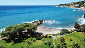 Un paysage côtier comprenant une plage, une côte rocheuse et un sentier menant à une zone herbeuse bordée d’arbres.