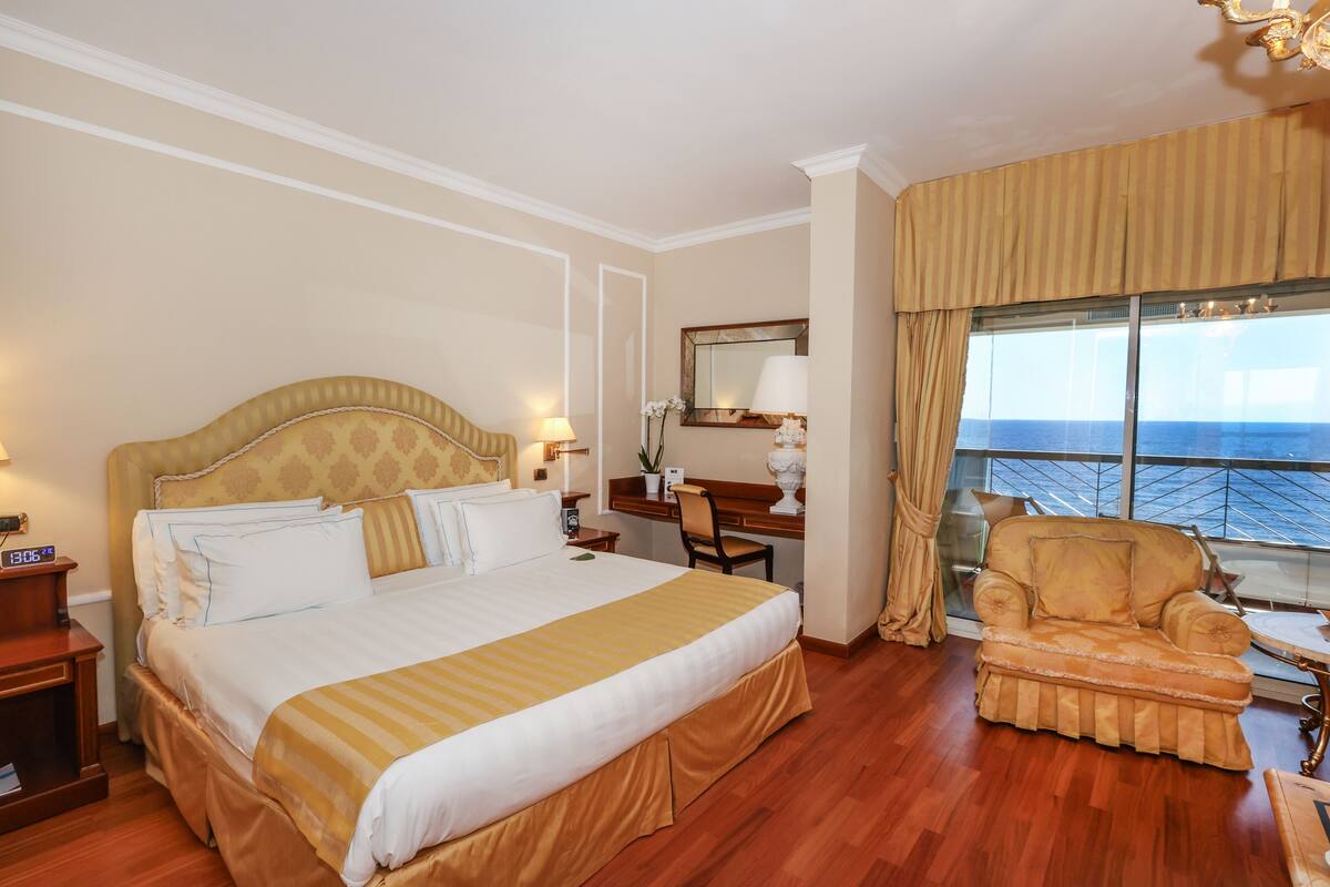 Ein Hotelzimmer mit einem großen Bett, einem Schreibtisch, einem Sessel und Blick aufs Meer.