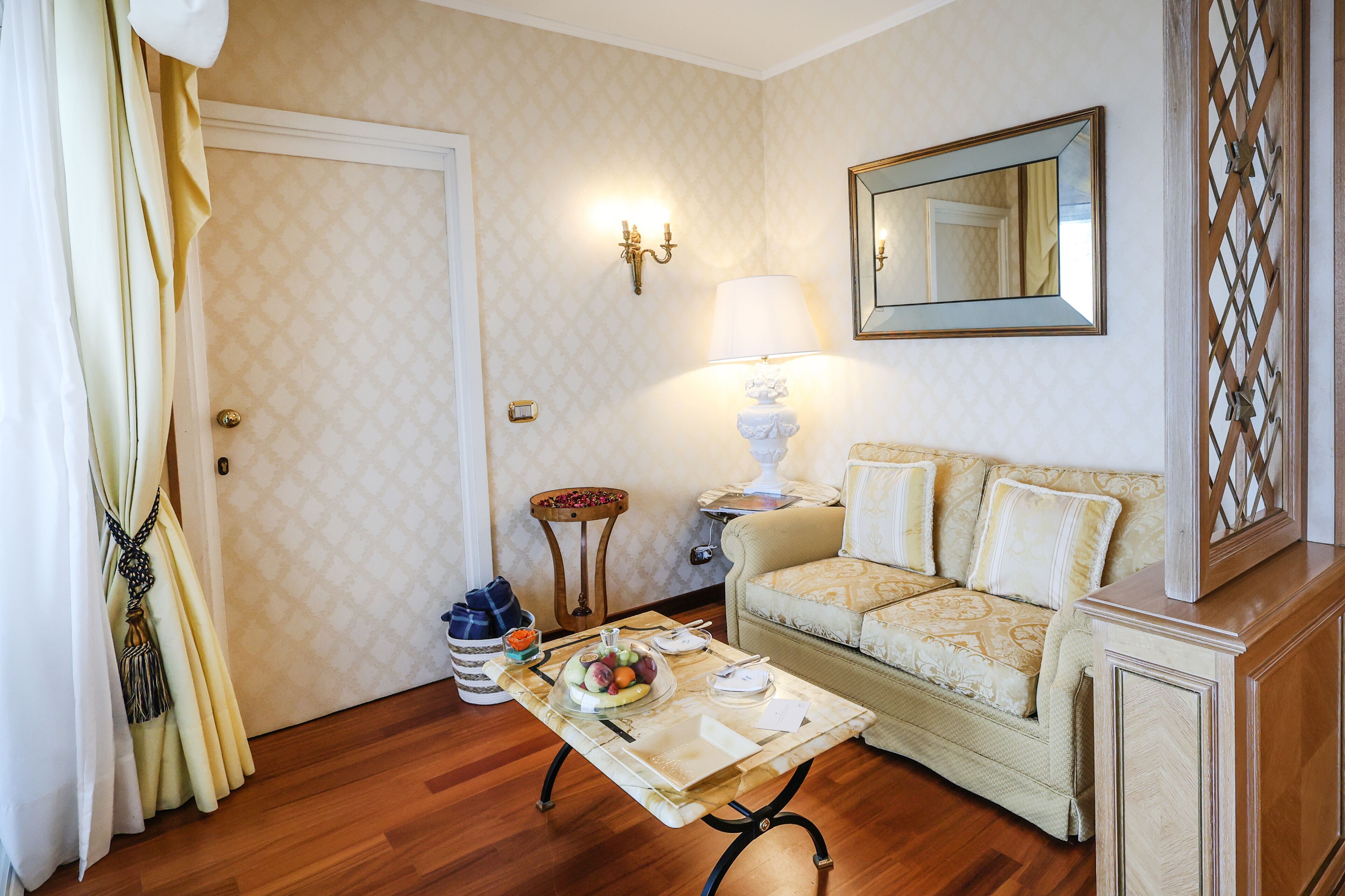 A room with a sofa, a table with a fruit bowl, a lamp, a mirror, and a door.