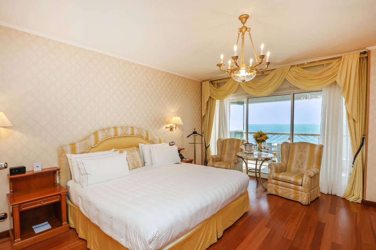 Ein Hotelzimmer mit einem großen Bett, einer Kristallleuchter, einem Sessel, einem Tisch mit einer Vase und Blick aufs Meer.