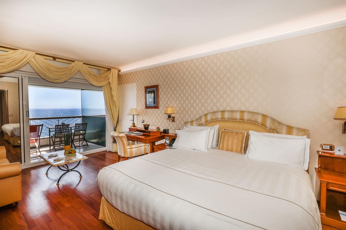 Ein Hotelzimmer mit einem großen Bett, einem Schreibtisch, einem Stuhl, einem kleinen Tisch und Meerblick durch eine Schiebetür.