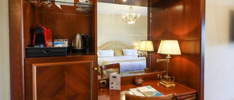 A hotel room with a wooden desk, a bed, a mirror, a chandelier, and a coffee maker.
