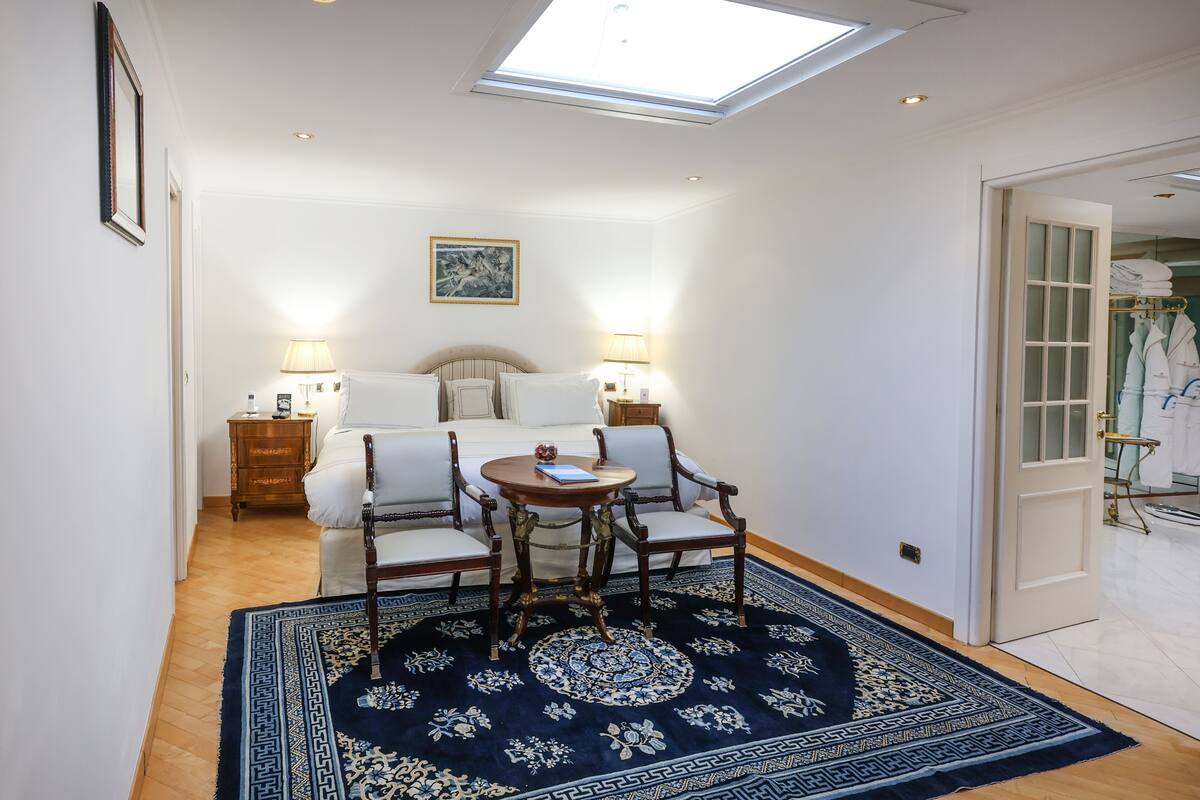 Ein Zimmer mit Dachfenster, einem Bett mit Kopfteil, zwei Nachttischen mit Lampen, einem runden Tisch mit Stühlen und einem gemusterten Teppich.