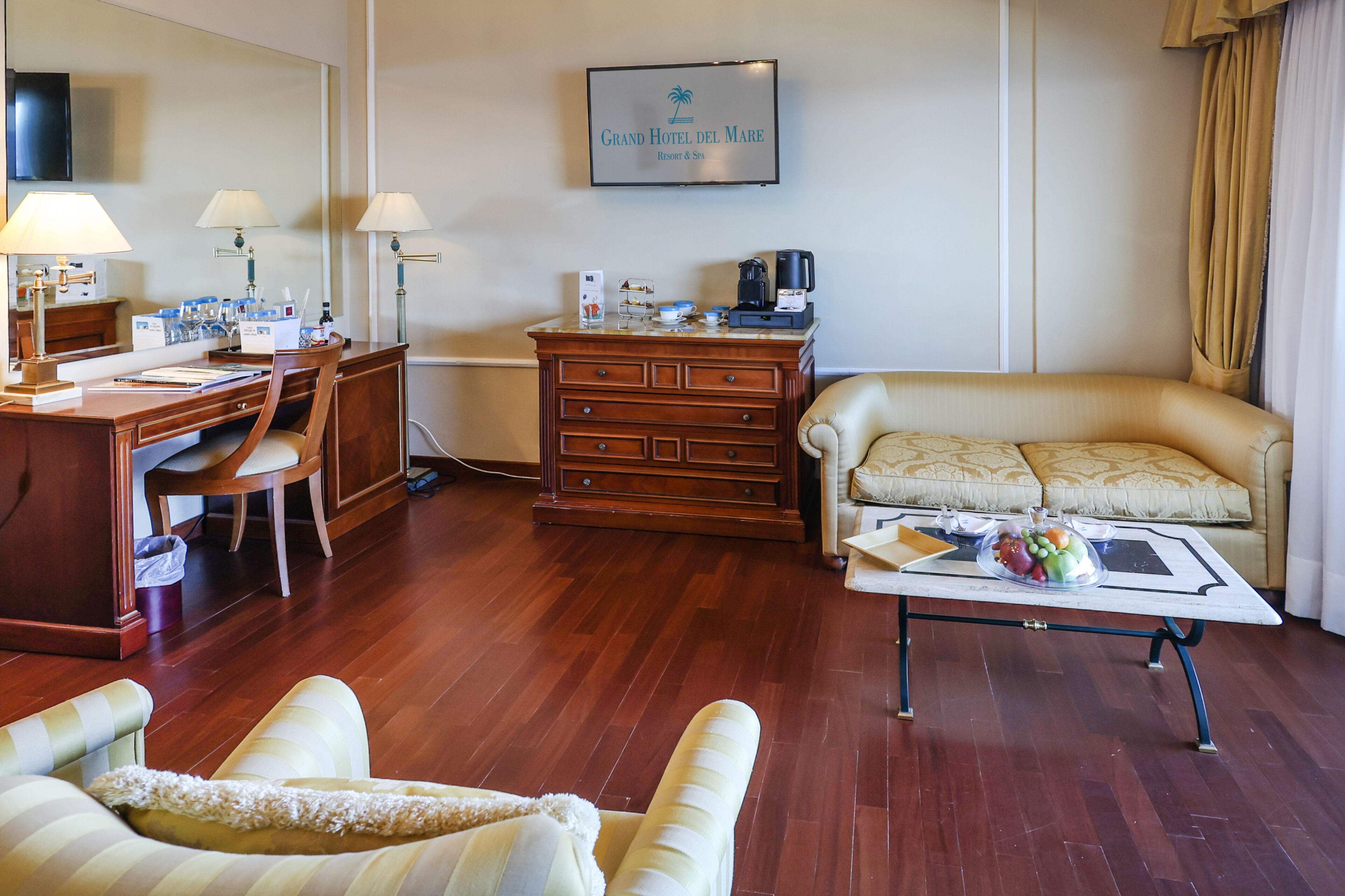 A hotel room with a wooden desk, a sofa, a coffee maker, and a framed sign on the wall.
