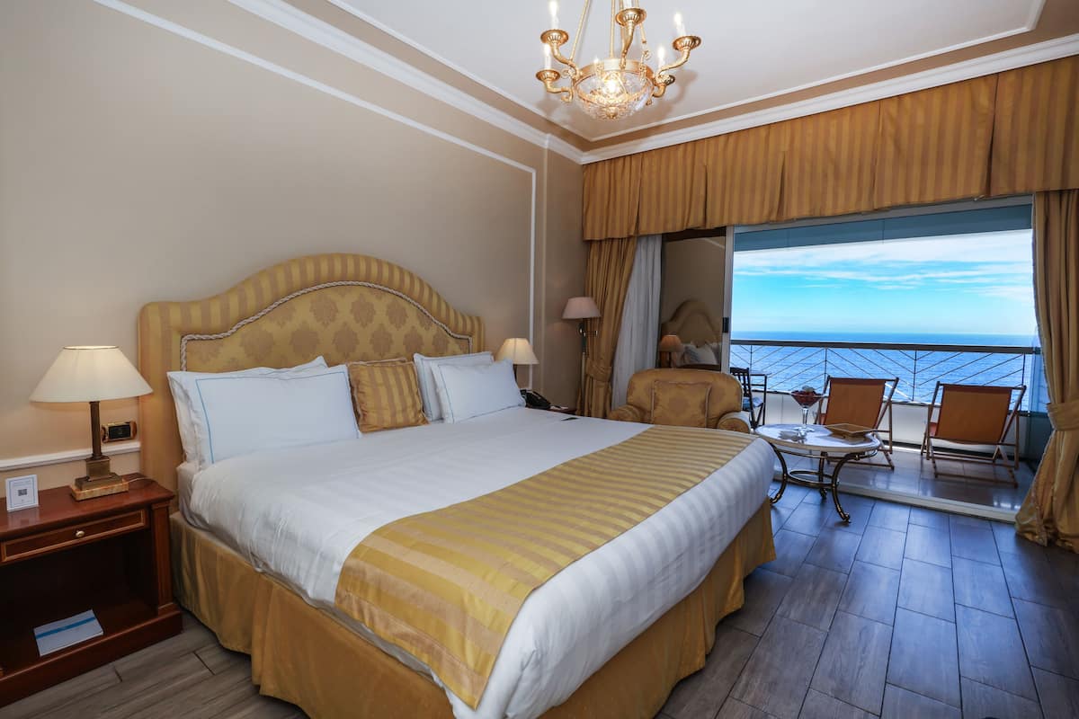 Ein Hotelzimmer mit einem großen Bett, einer Sitzecke mit Tisch und Stühlen, einer Kristallleuchtre und einem Balkon mit Blick.