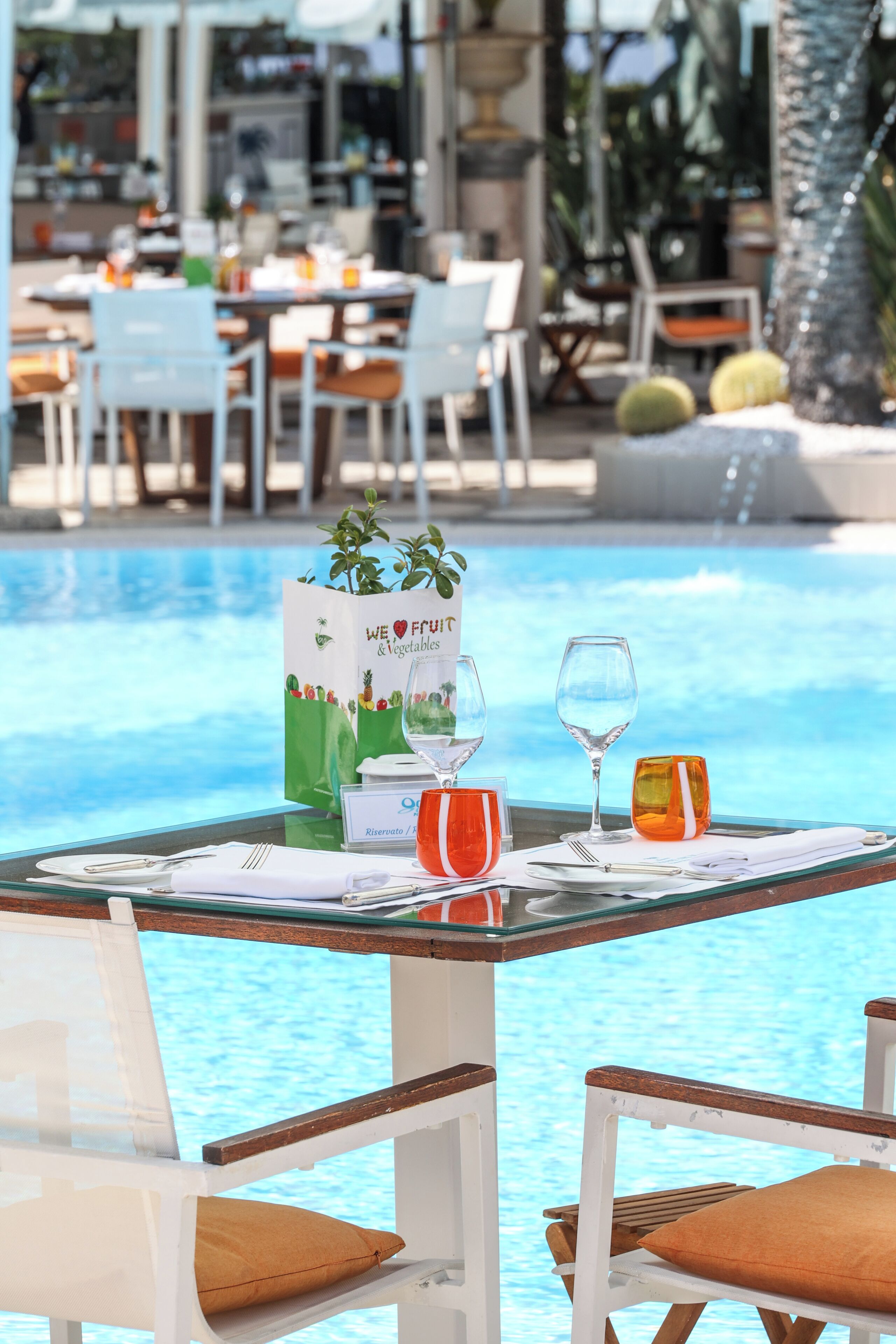 Un área de comedor junto a la piscina con mesas preparadas para dos, un atril con la carta y vistas despejadas a la piscina.