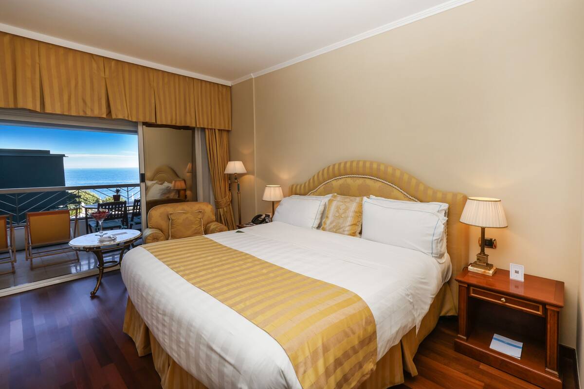 Ein Hotelzimmer mit einem großen Bett, zwei Nachttischen mit Lampen, einem Sessel, einem kleinen Tisch und Blick aufs Meer.