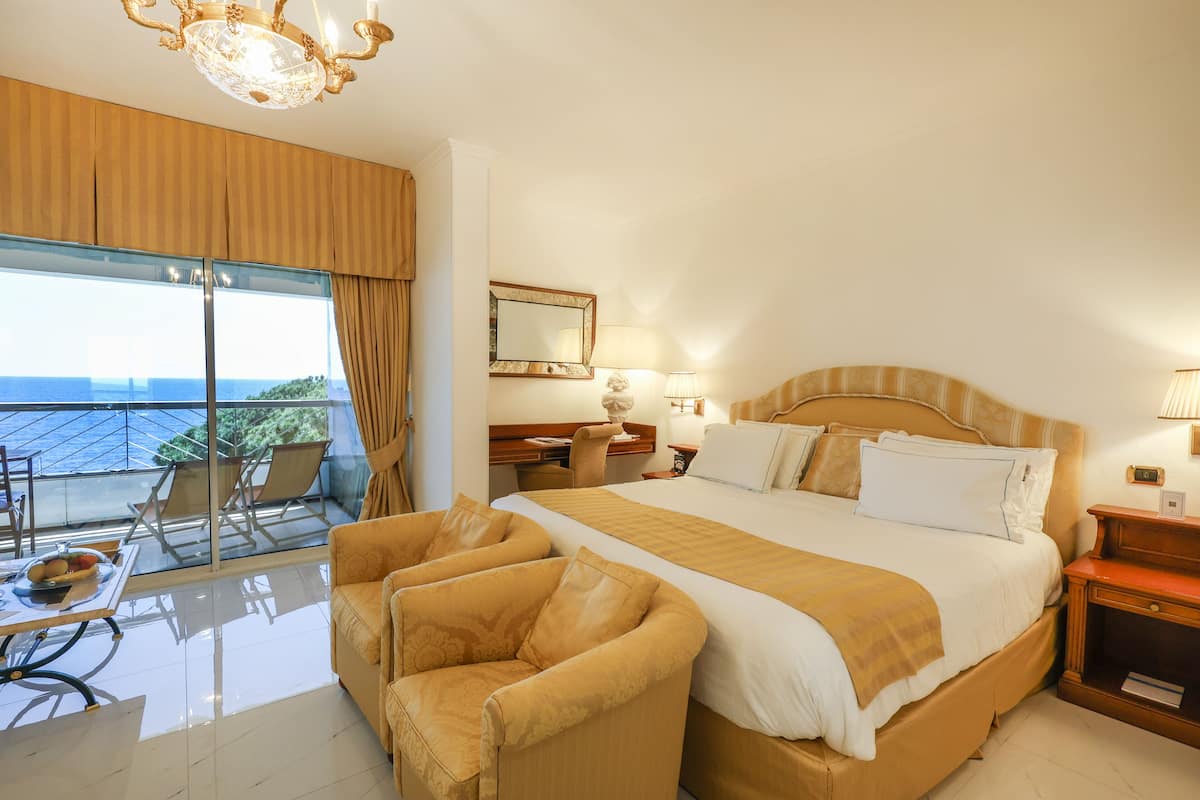 Ein Hotelzimmer mit einem großen Bett, einem Schreibtisch, einem Sessel, einem Balkon mit Blick und einer Kristallleuchter.
