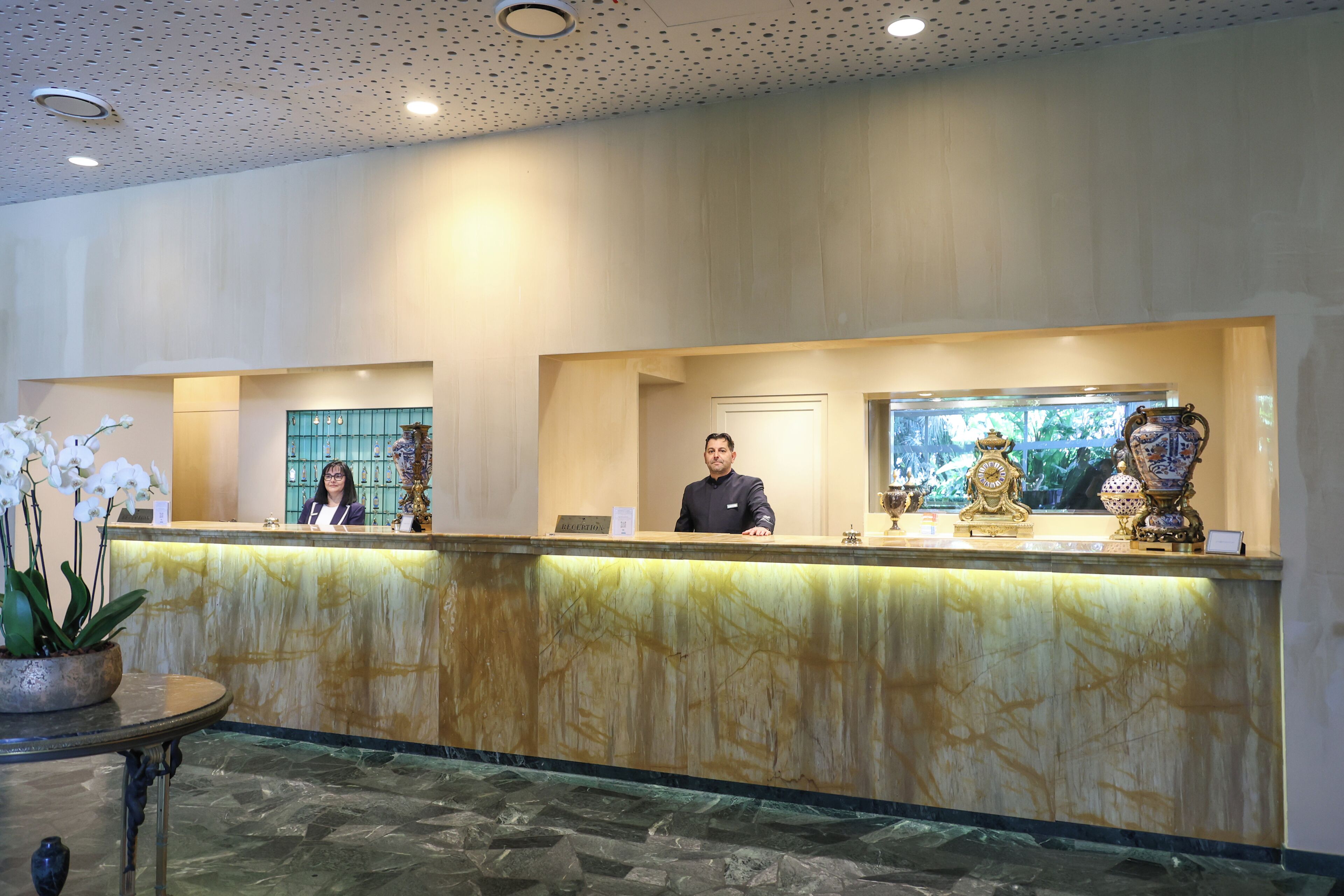 A hotel reception area with a marble counter, two receptionists, and decorative vases.