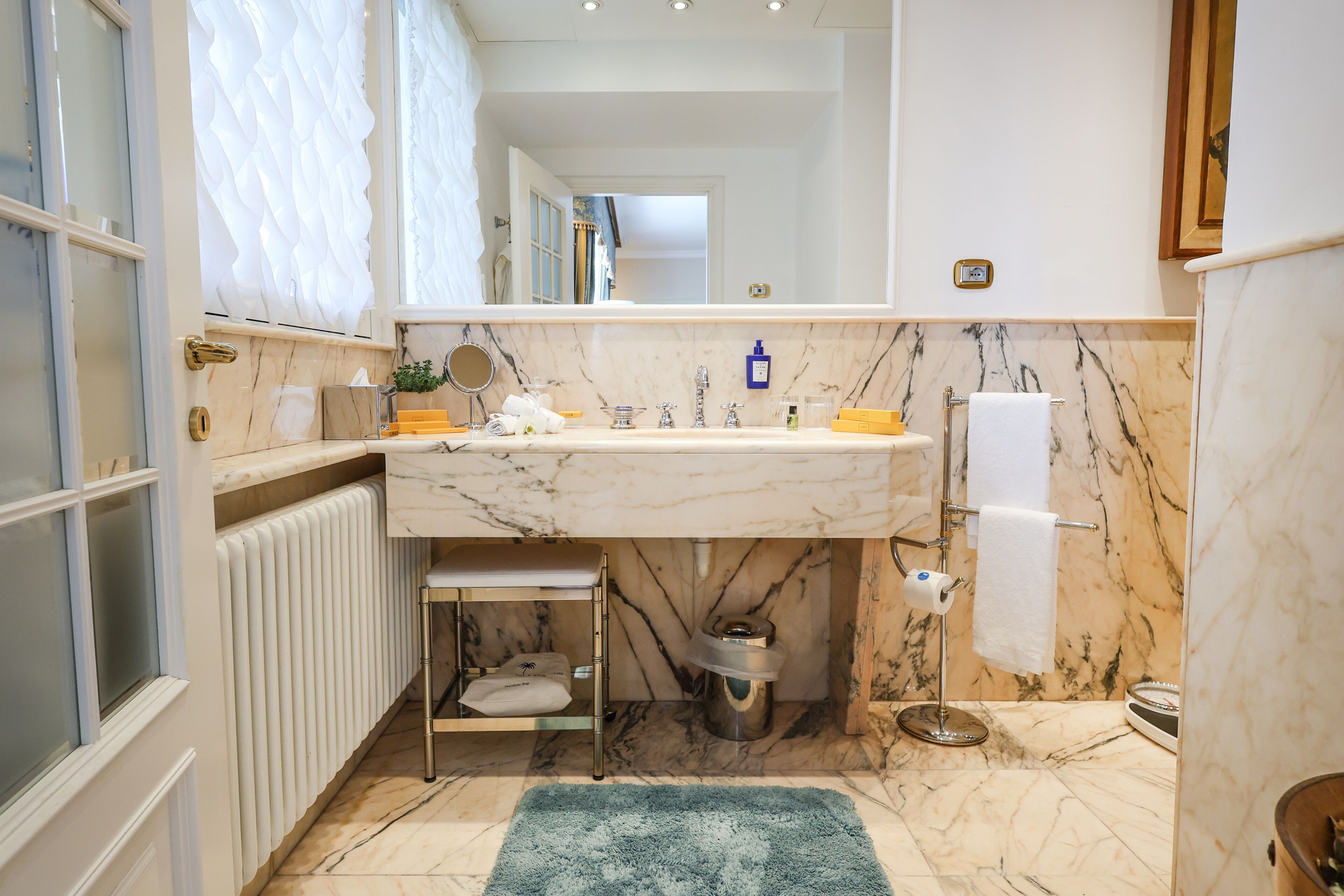 Un baño con lavamanos de mármol, toallero, espejo, radiador y una alfombra azul.