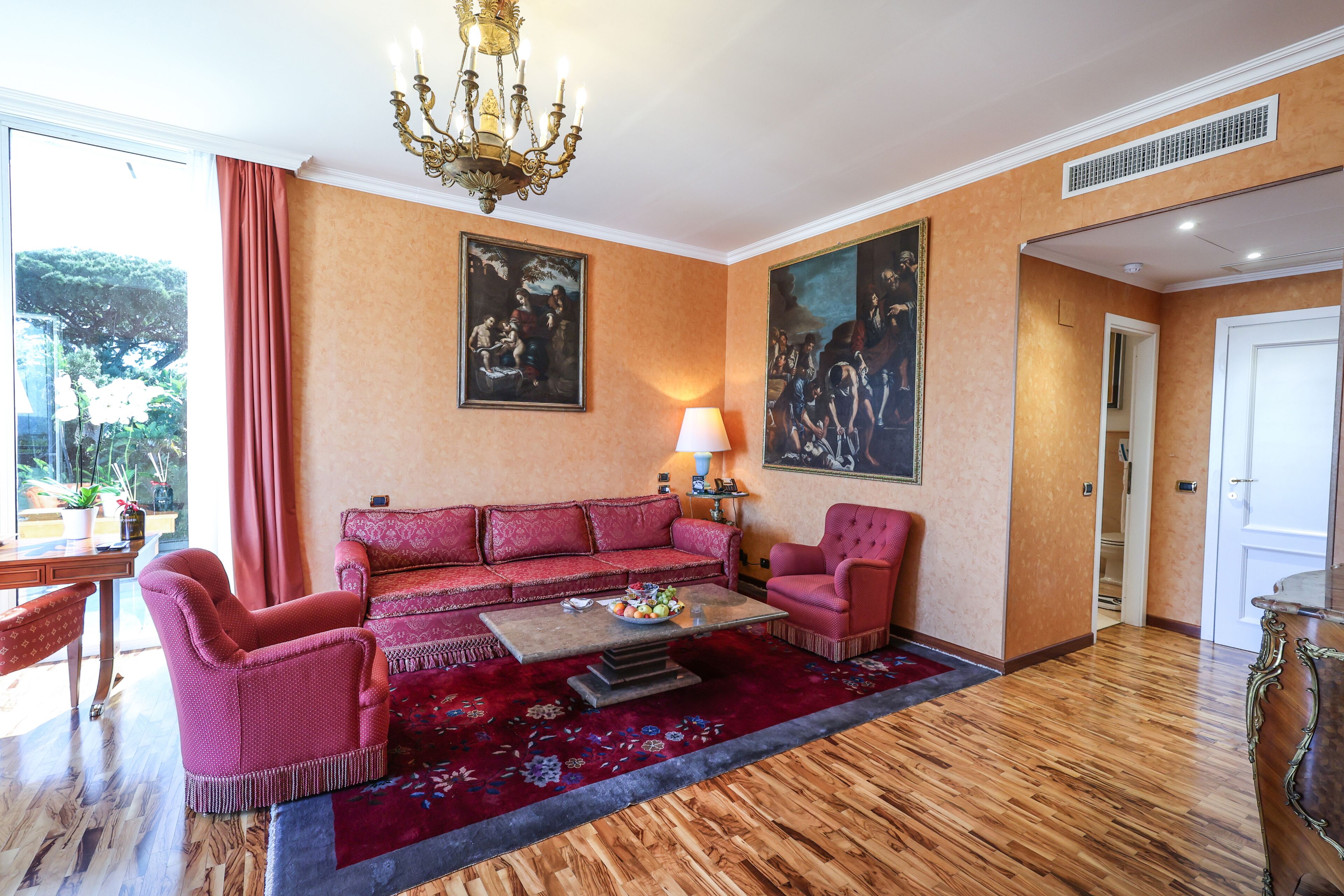A room with a red sofa set, a coffee table, a chandelier, and two paintings on the wall.