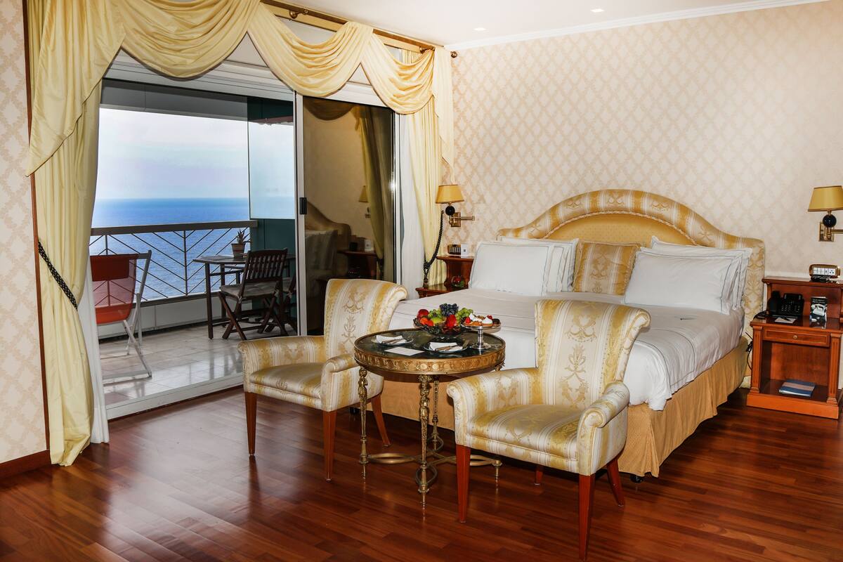 Ein Hotelzimmer mit einem Bett, zwei Sessel, einem kleinen runden Tisch, einem Nachttisch und Blick aufs Meer.