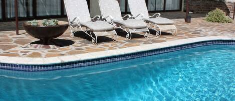 A swimming pool with clear blue water, surrounded by lounge chairs and a stone-tiled deck.