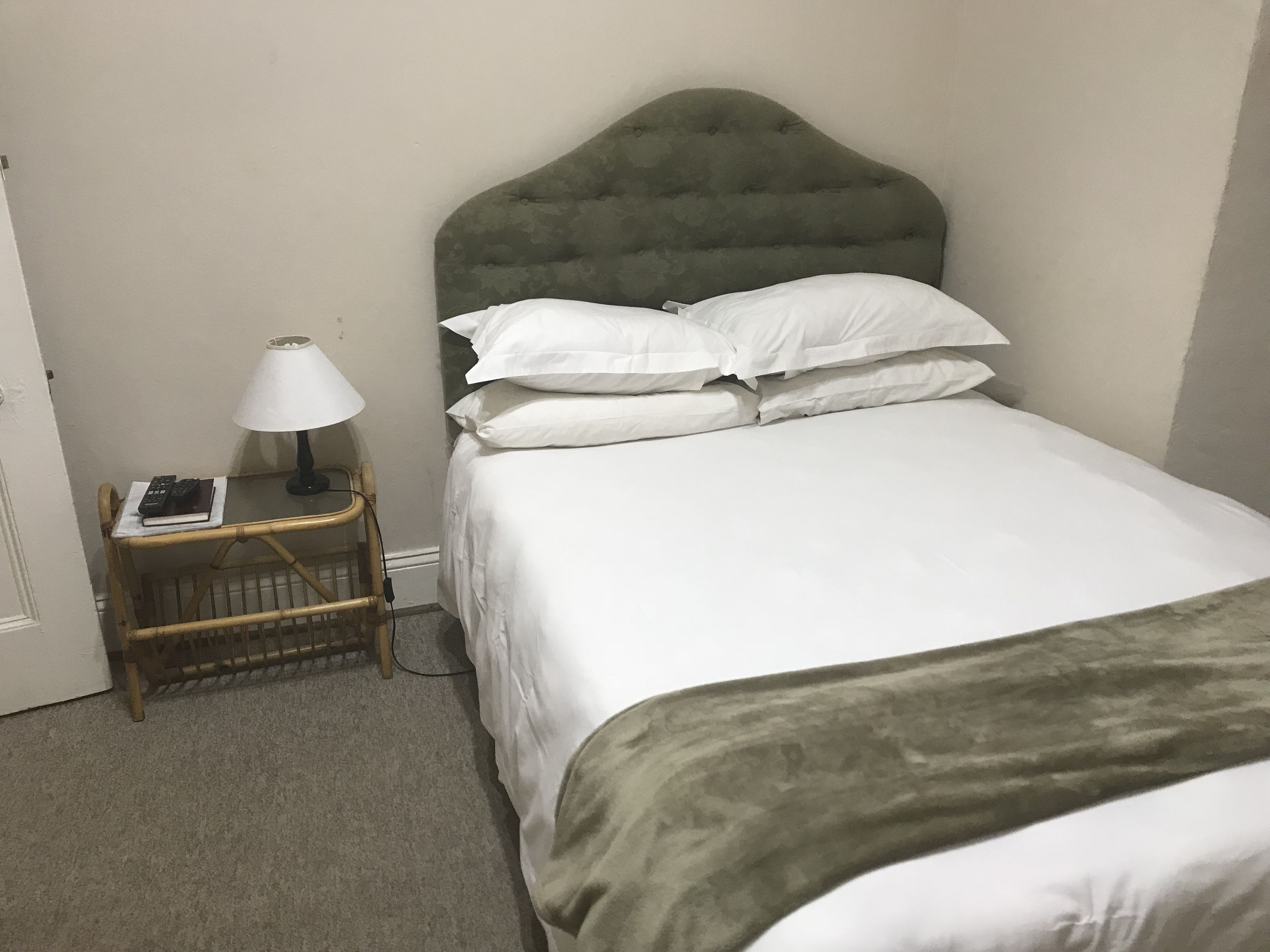 Een netjes opgemaakt bed met witte lakens en een groen hoofdeinde, een nachtkastje met een lamp en een boek.