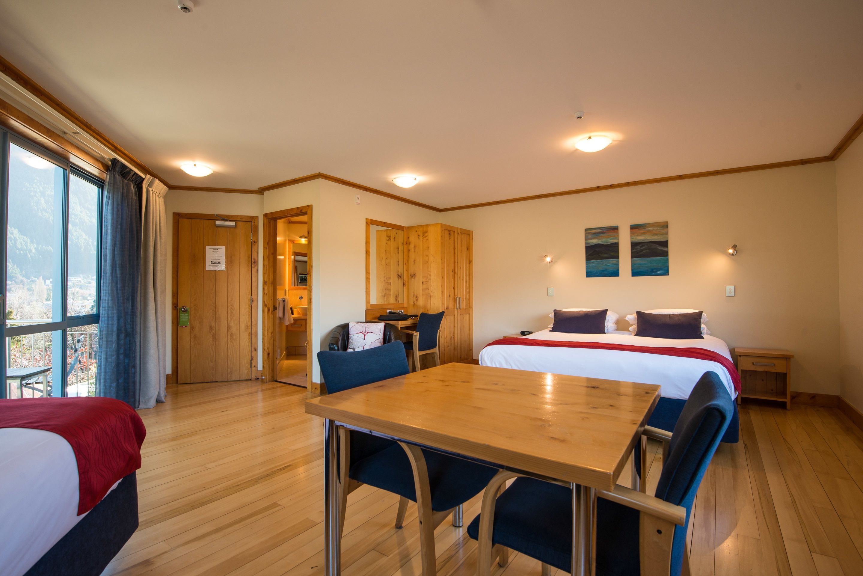 A hotel room with a wooden floor, a bed with a red blanket, a dining table with chairs, a wooden wardrobe, and a window with curtains.
