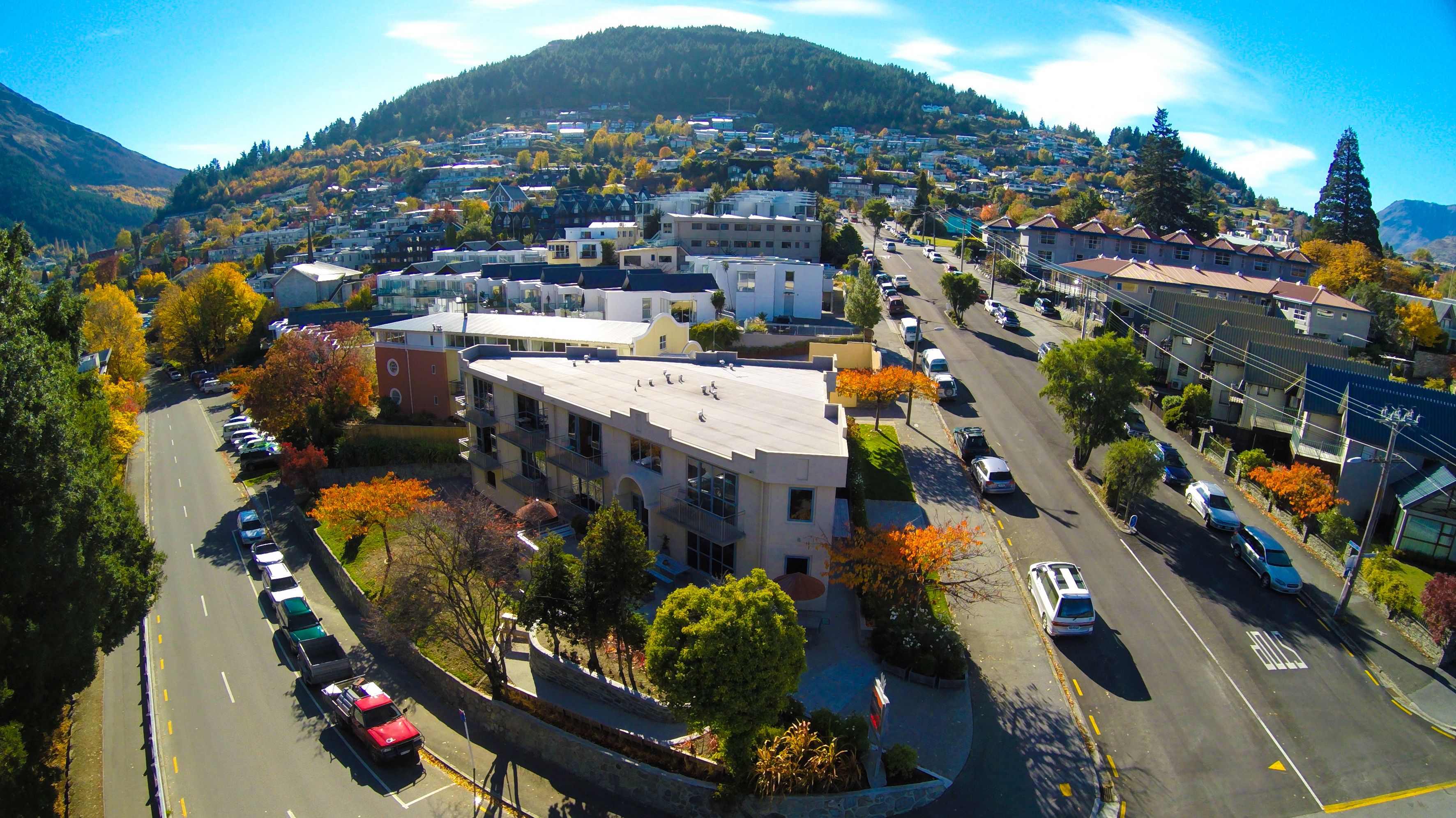 An aerial view of a town with a mix of residential and commercial buildings, a main road, and a hilly backdrop.
