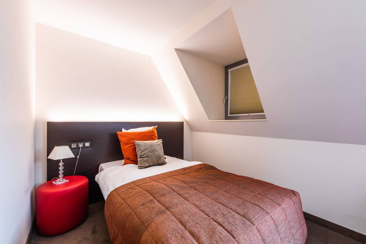 Un dormitorio con cama, un reposapiés rojo, una mesita de noche con lámpara, una ventana y un techo inclinado.
