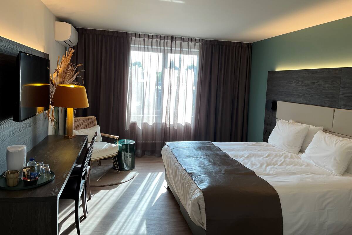 Habitación de hotel con cama, escritorio con lámpara, televisor y ventana con cortinas.