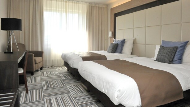 A hotel room with two beds, a desk, a chair, and a large window with curtains. - Malpertuus (Riemst)