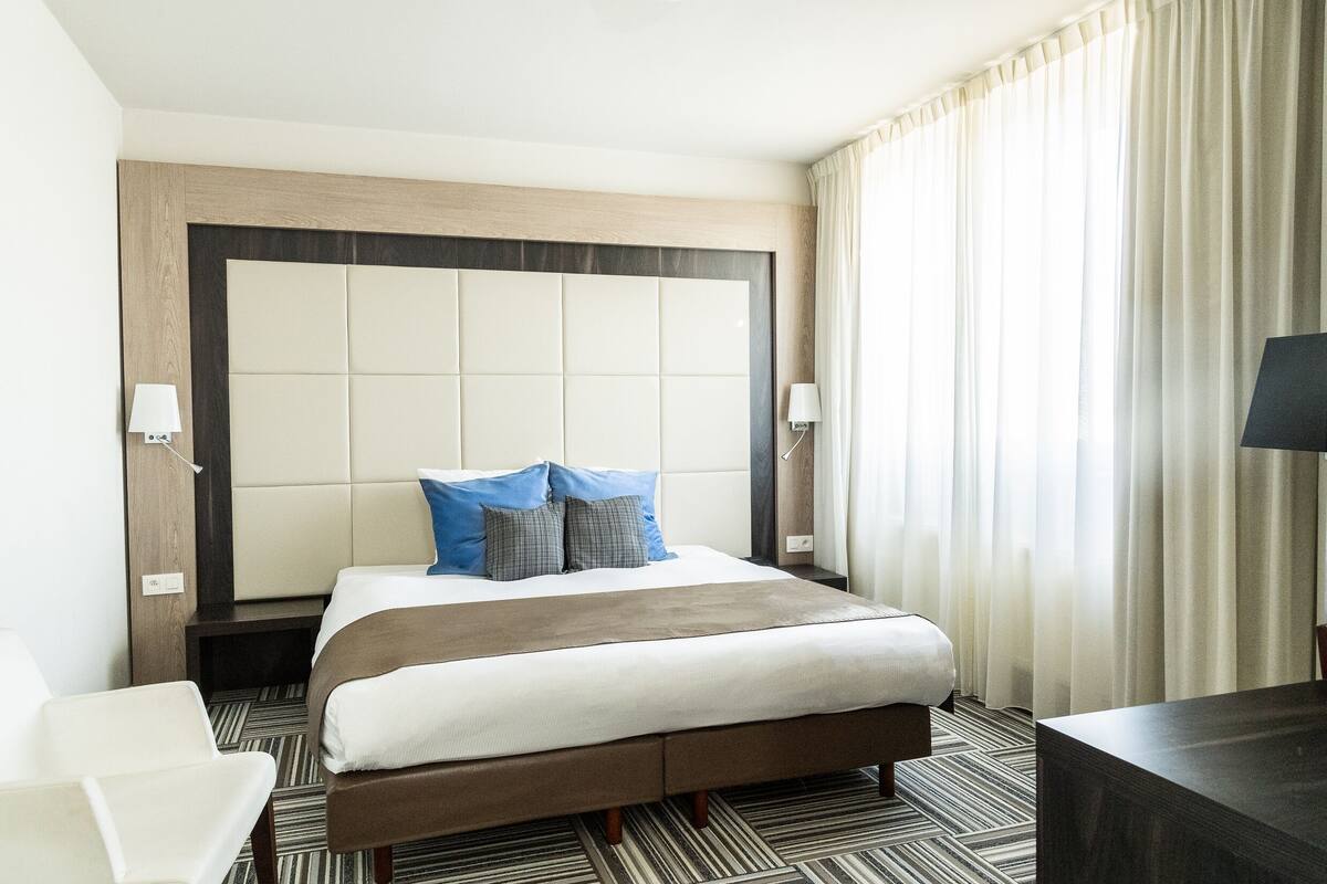 Habitación de hotel con una cama grande, una mesita de noche, una silla y vistas al exterior a través de la ventana.