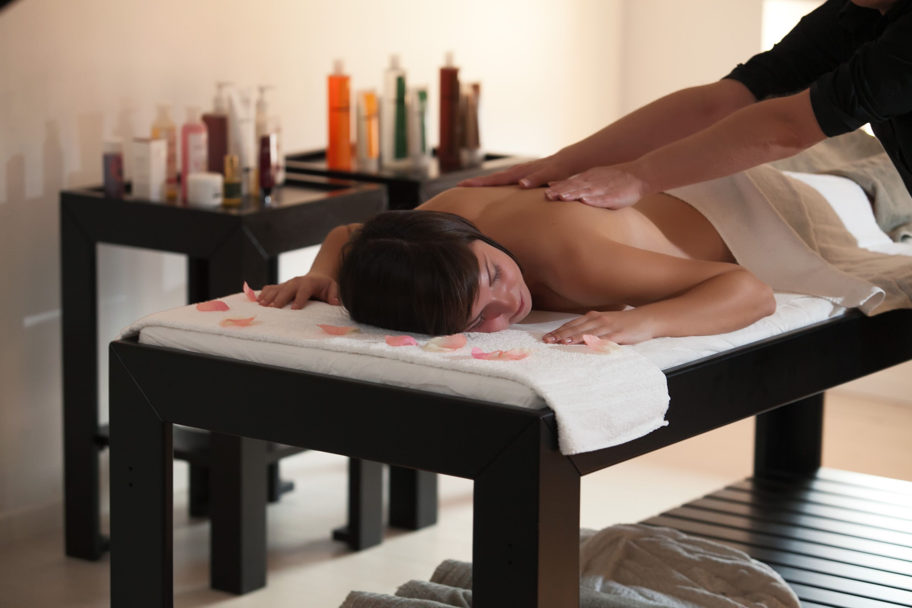 A massage table with a towel and rose petals, a massage therapist's hands on a client's back, and bottles of massage oils on a table in the background.