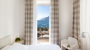 A hotel room with a bed, a small table with a flower, a chair, and a view of the sea and mountains through a window. - Hotel Lago di Garda (Nago-Torbole)
