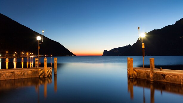 A calm waterfront at dusk with a pier extending into the water, surrounded by mountains. - Hotel Lago di Garda (Nago-Torbole)