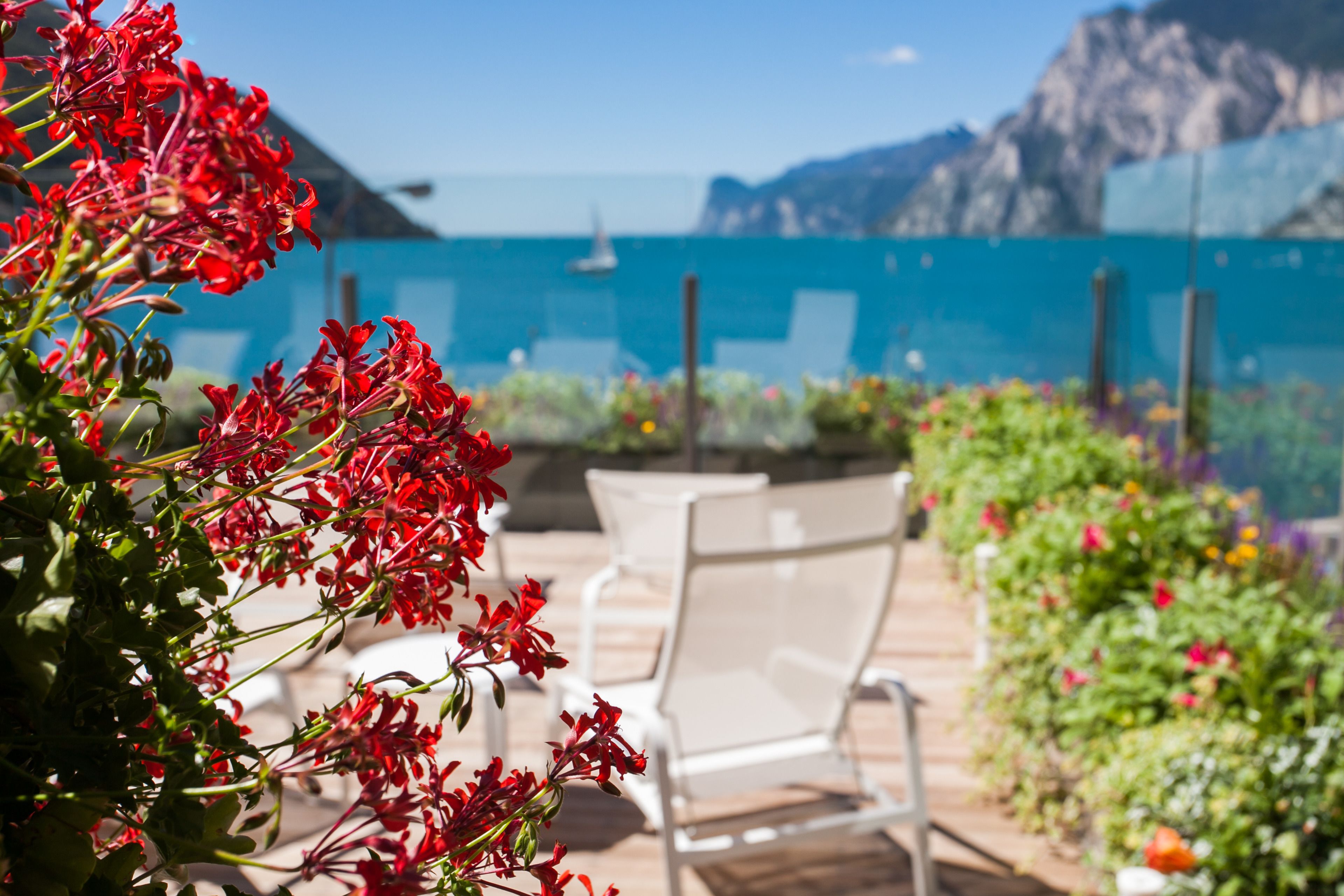 Un patio con sillas blancas, flores rojas y vistas al mar y a las montañas.