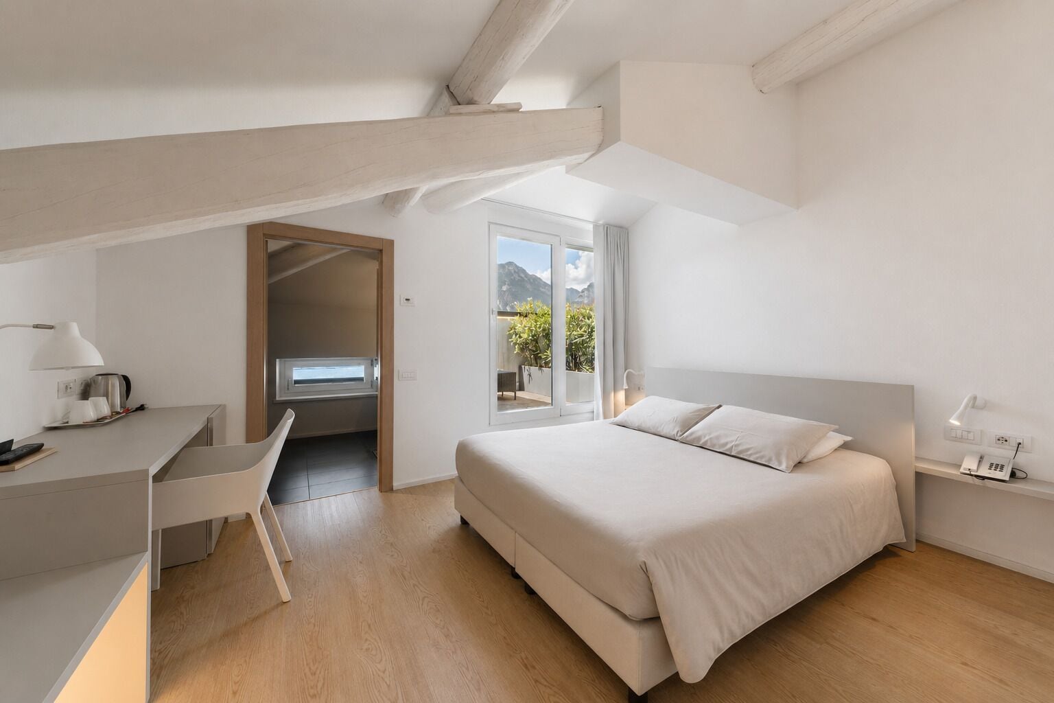 A modern bedroom with a bed, desk, and a view of mountains through a large window.