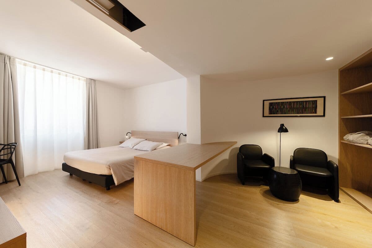 Moderni hotellihuone, jossa on sänky, työpöytä, kaksi tuolia, lamppu ja kirjahylly.