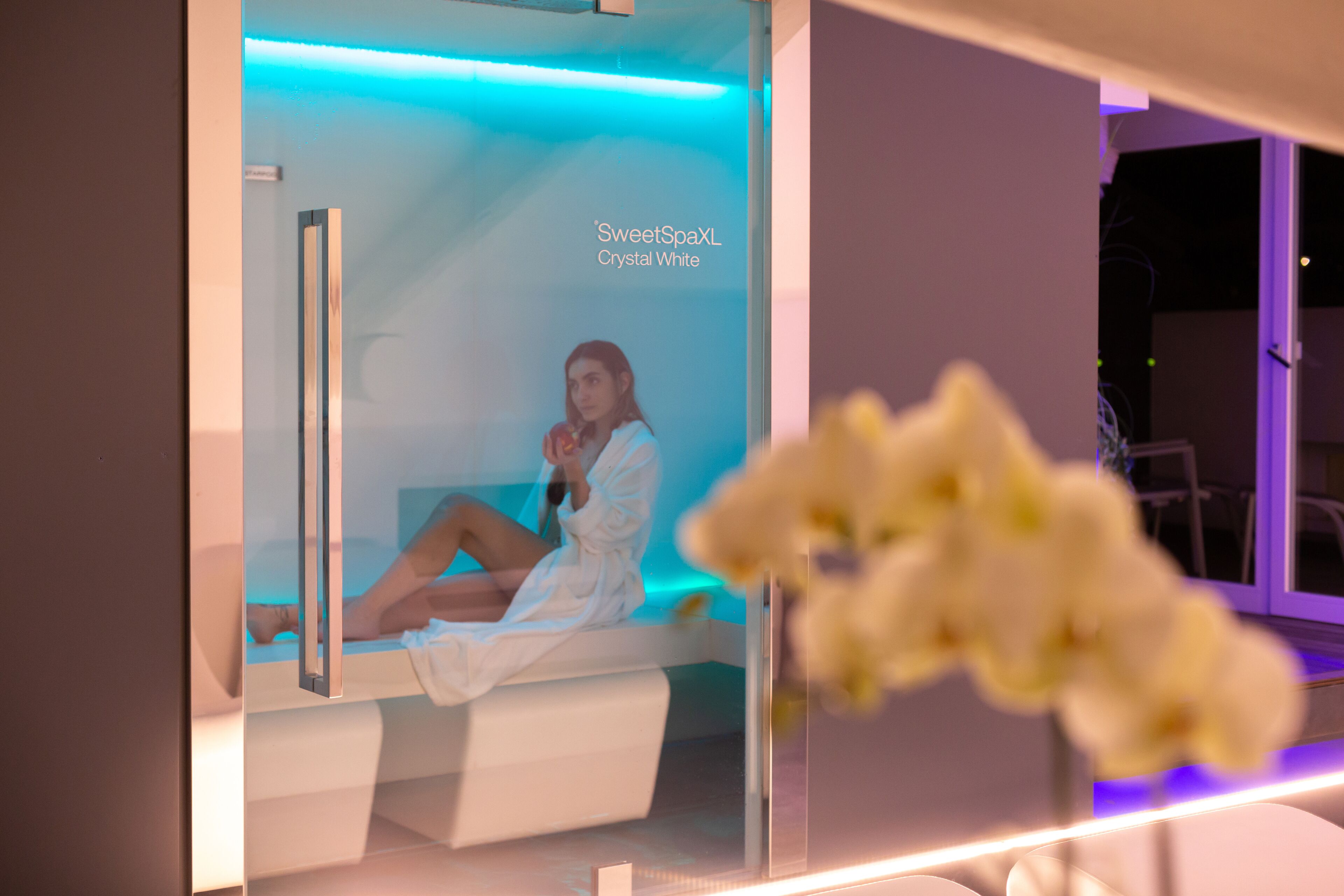 A spa room with a glass partition, a person in a robe, and a sign indicating 'SweetSpaXL Crystal White'.