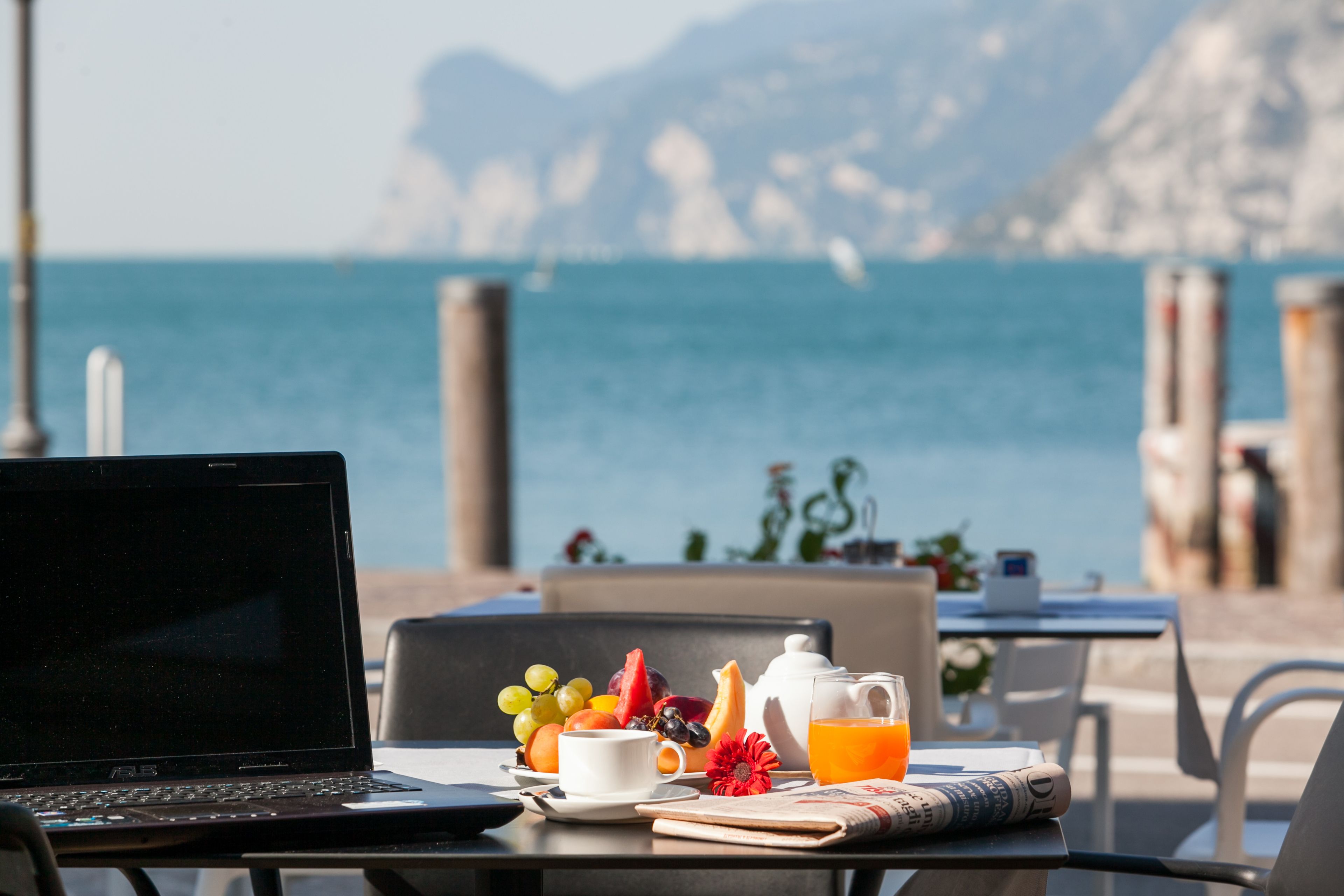 A laptop on a table with a breakfast setting including fruits, a teapot, and a glass of orange juice, overlooking the sea.