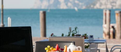 A laptop on a table with a breakfast setting including fruits, a teapot, and a glass of orange juice, overlooking the sea.