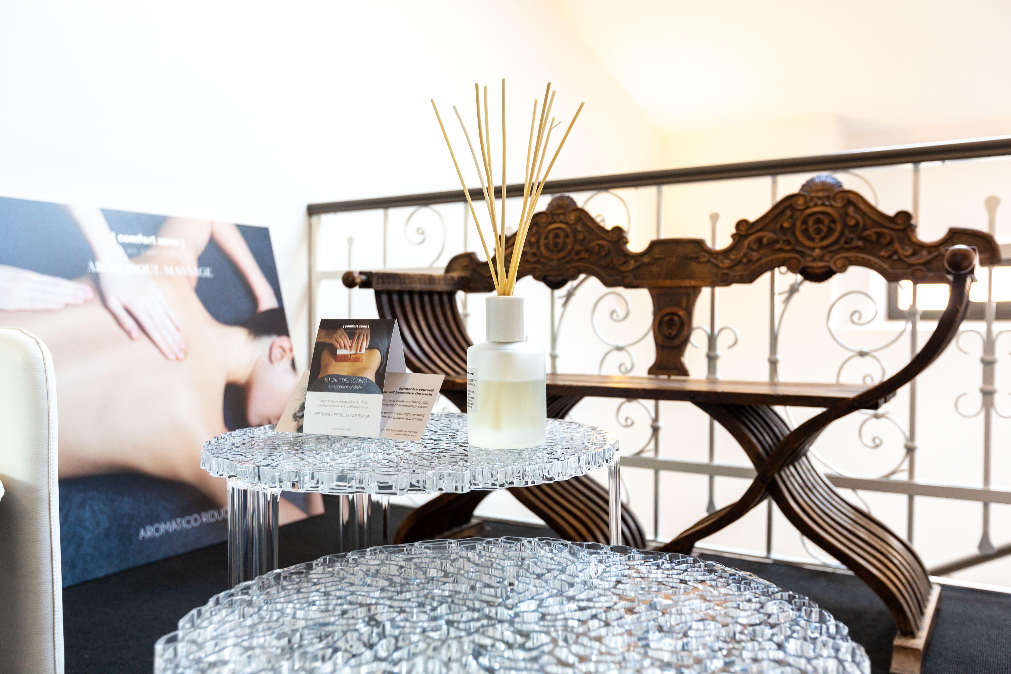 A glass table with a bottle and reed sticks, a framed picture, and a decorative chair.